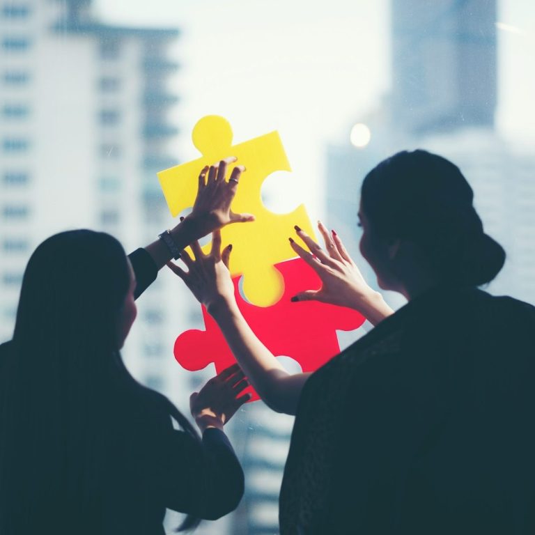 Zwei Personen fügen farbige Puzzlestücke zusammen vor einem modernen Gebäude.